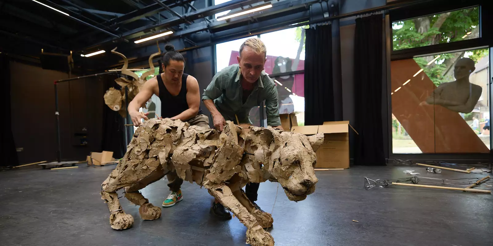 Two people operate a large, detailed animal puppet made from cardboard in a studio space.