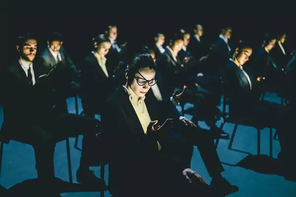 A still from Figures in Extinction [2.0] showing performers sat on chairs, wearing glasses and suits and looking at their phones