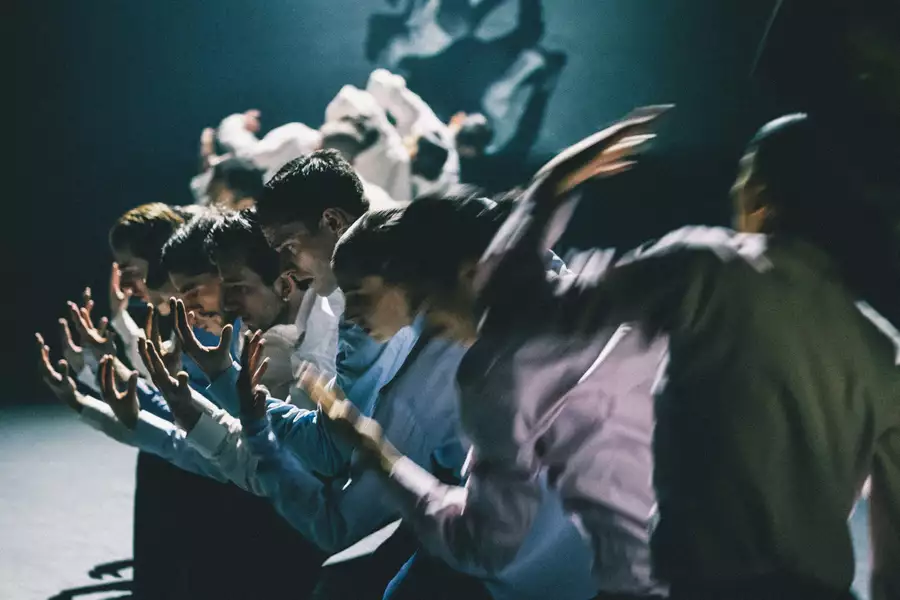 A group of dancers stood in a line with their arms extended forward, moving in blurred motion. The lighting is dim, casting dramatic shadows.