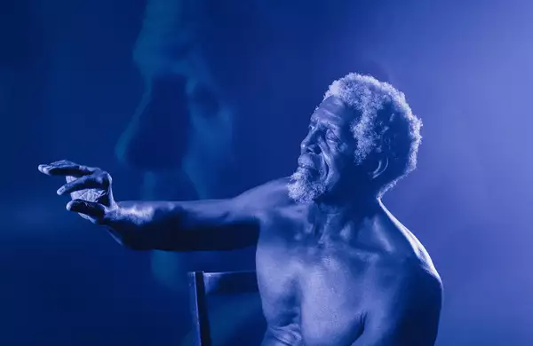 A shirtless elderly man with curly white hair and a beard sits on a chair, reaching out with an expressive hand. The image has a blue monochromatic tone with a soft-focus overlay of his face in the background.