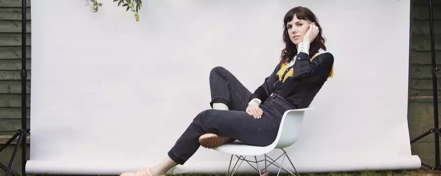 Anna Meredith sits in a modern white rocking chair outdoors, posing in front of a white backdrop with green branches hanging above. She wears a black shirt with yellow detailing, dark jeans, and peach sneakers.