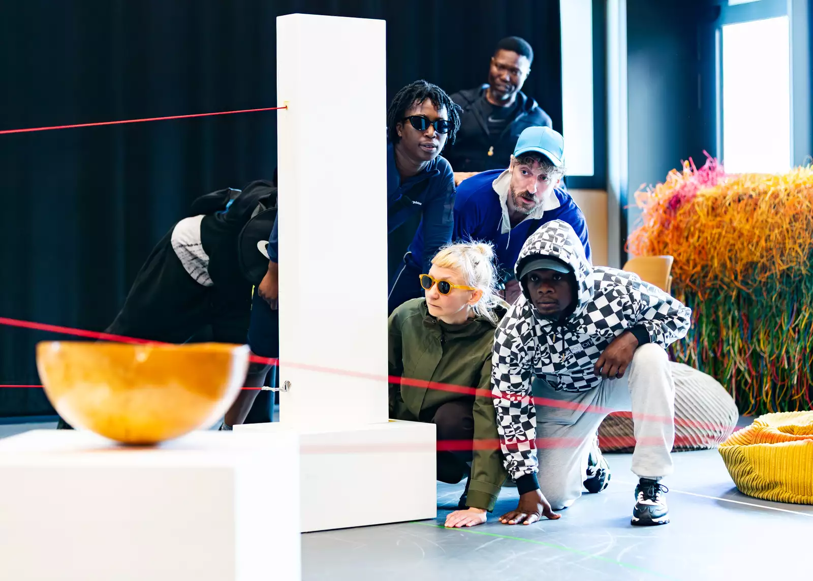 Five people hide behind a plinth and lasers in the rehearsal room, looking at a bowl