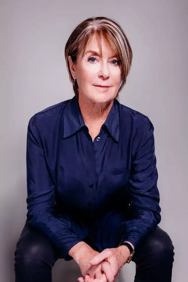 SUZANNE LACY sitting down wearing a dark blue shirt, looking at the camera. The background is grey.