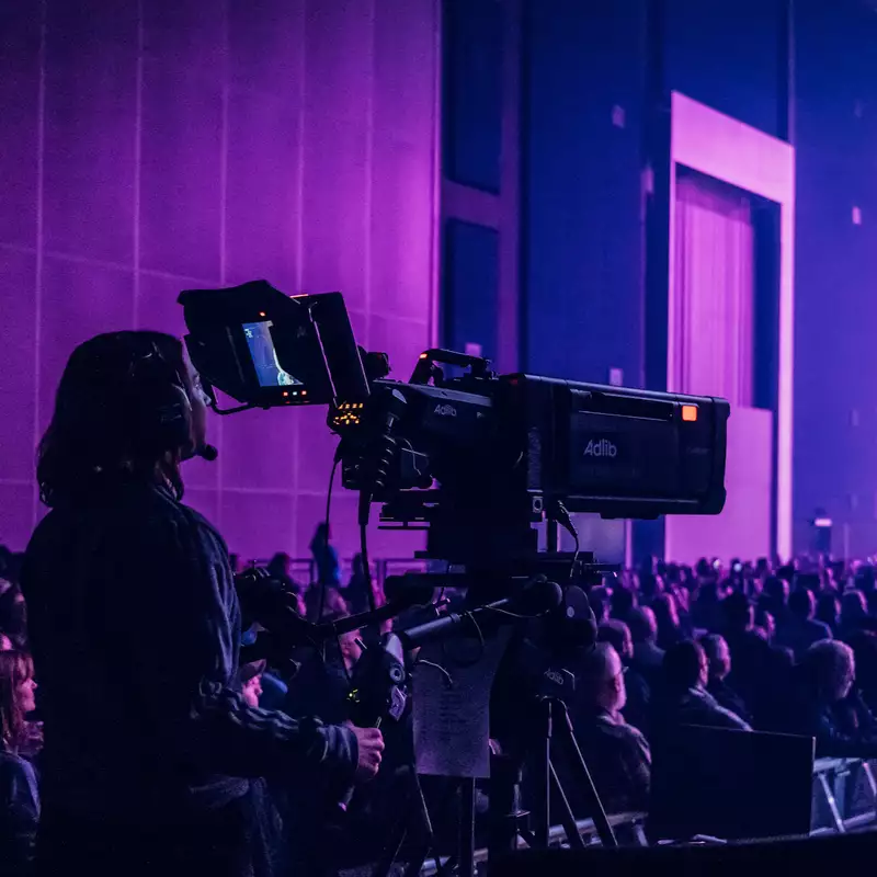A camera operator films a live event in the Warehouse, which is filled with people. There is dramatic purple lighting with multiple cameras and bright stage beams.