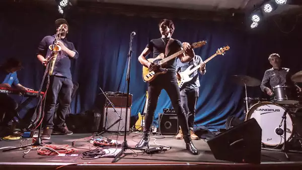 A five-member band performs on stage; one plays keyboard, one plays saxophone, one sings and plays electric guitar, one plays bass, and another plays drums, all set against a blue curtain backdrop with stage lights above.