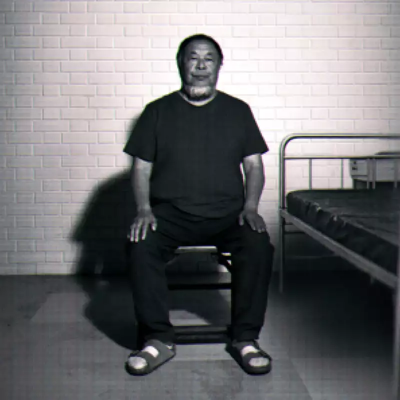 Black-and-white image of Ai Weiwei sitting on a chair beside a simple metal bed against a brick wall.