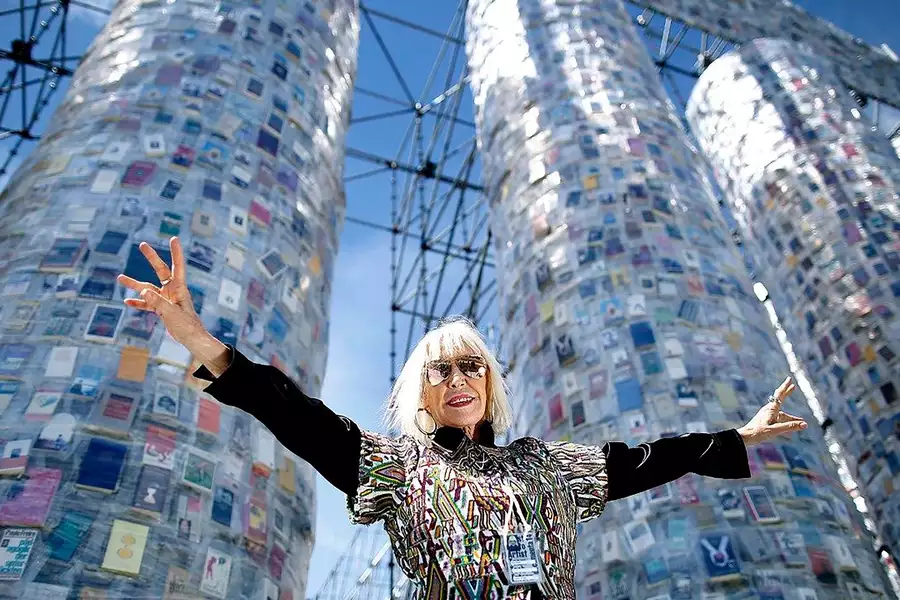 A photograph of Marta Minujín, stood with her arms outstretched in front of 3 large pillars wrapped in books and cling-film.