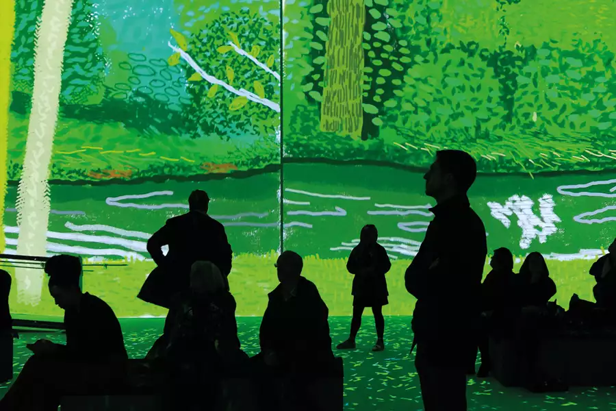 People sit and stand in David Hockney: Bigger & Closer (not smaller & further away) at Aviva Studios. Hockney's image of a forest is projected on the walls.