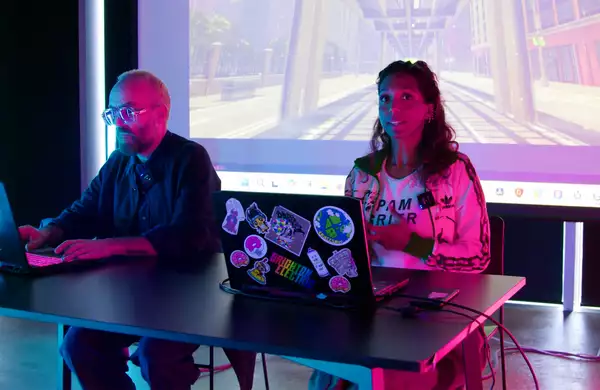 Simon Wilkinson and Myra Appannah sat at a desk on their computers. Behind them is a big screen showing a game.