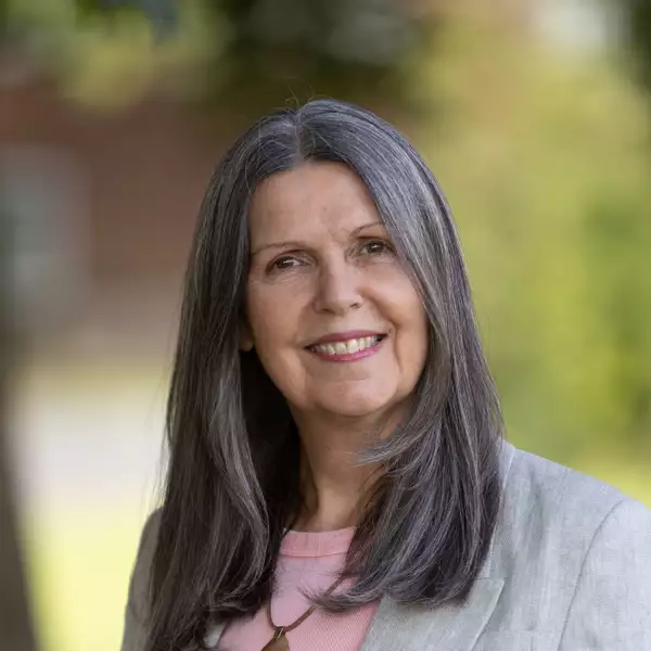 Headshot of Marie Greenhalgh smiling and wearing a light pink top and grey blazer. They are stood outside and the background is blurred.