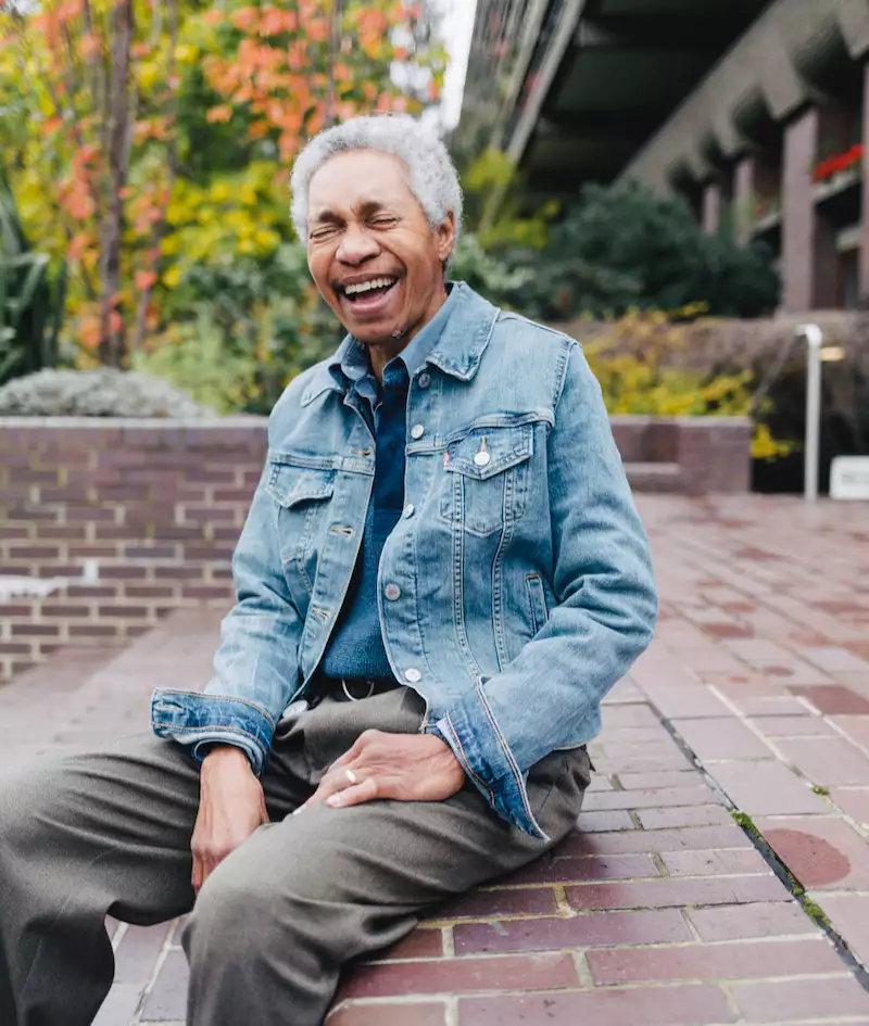 Glenn laughing and wearing a denim jacket and grey trousers. They are sat outside on a tiled platform.