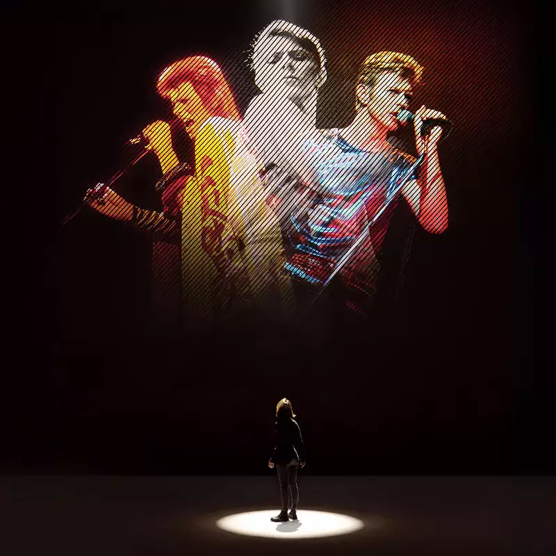 A person looking at five versions of David Bowie singing on a large screen