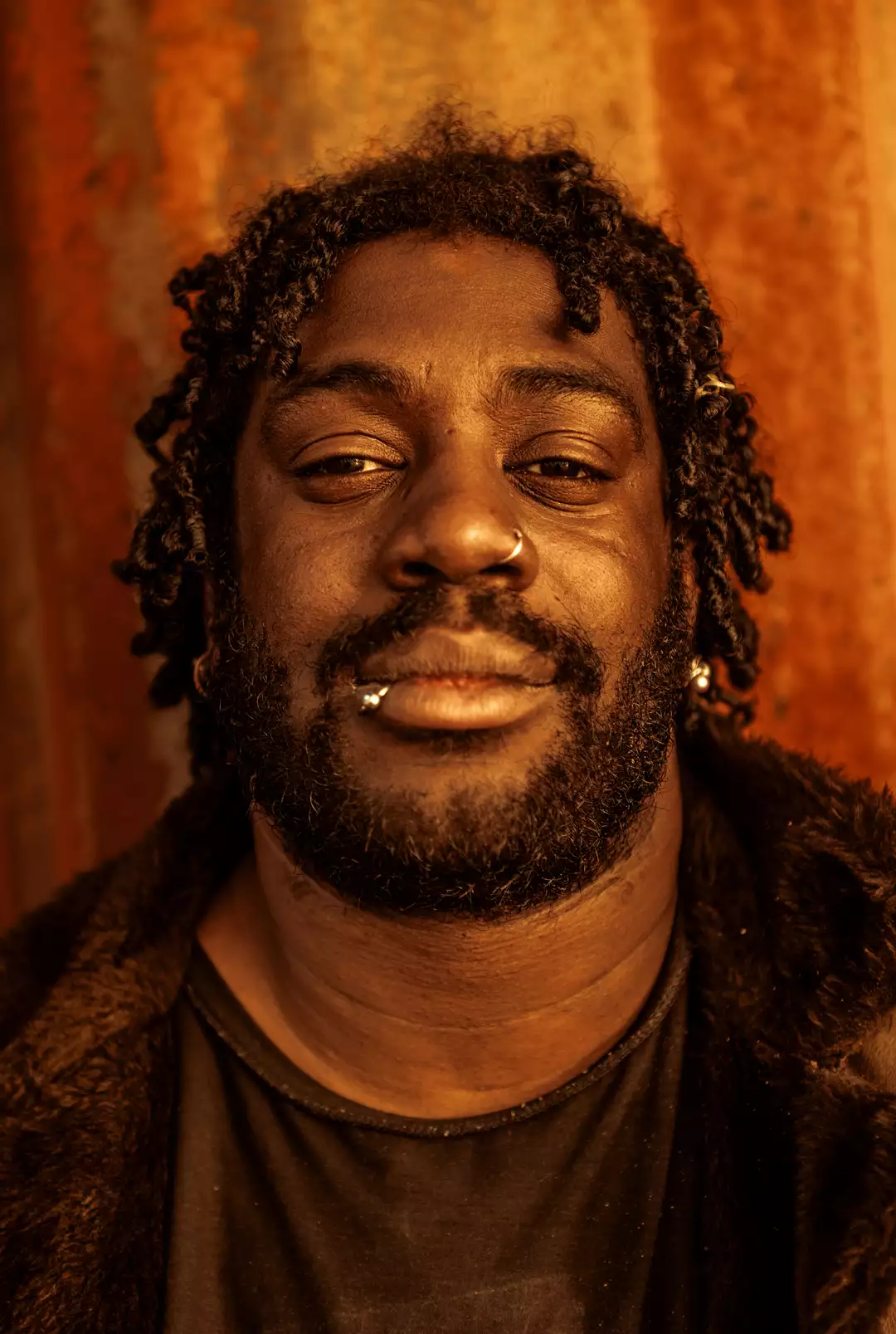 Close up of a face. The person is half smiling, looking directly into the camera with semi-closed eyes. They wear a nose piercing and lip ring. They have a beard and short locks.