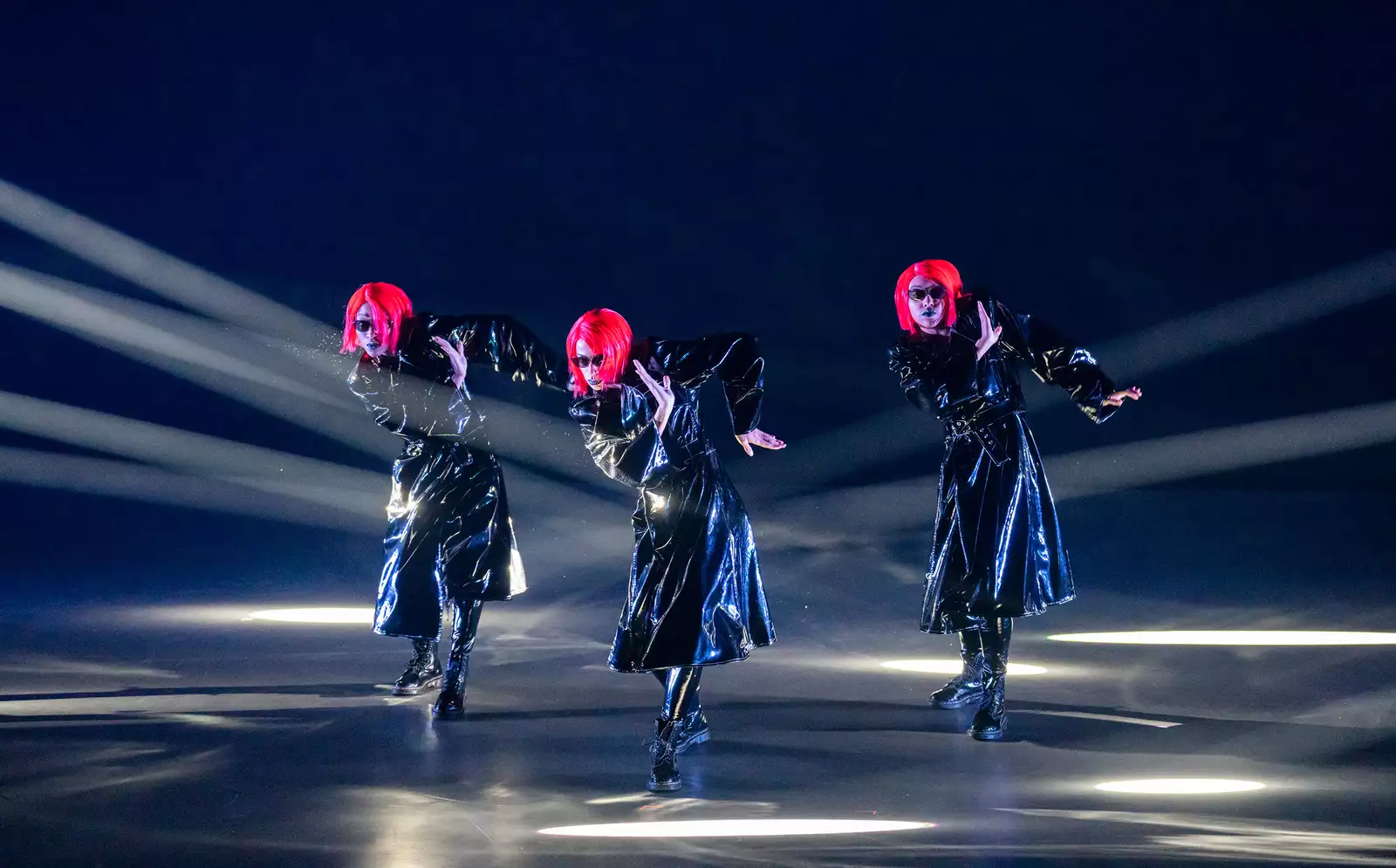 Three dancers on stage in Free Your Mind wearing long PVC coats and pink wigs