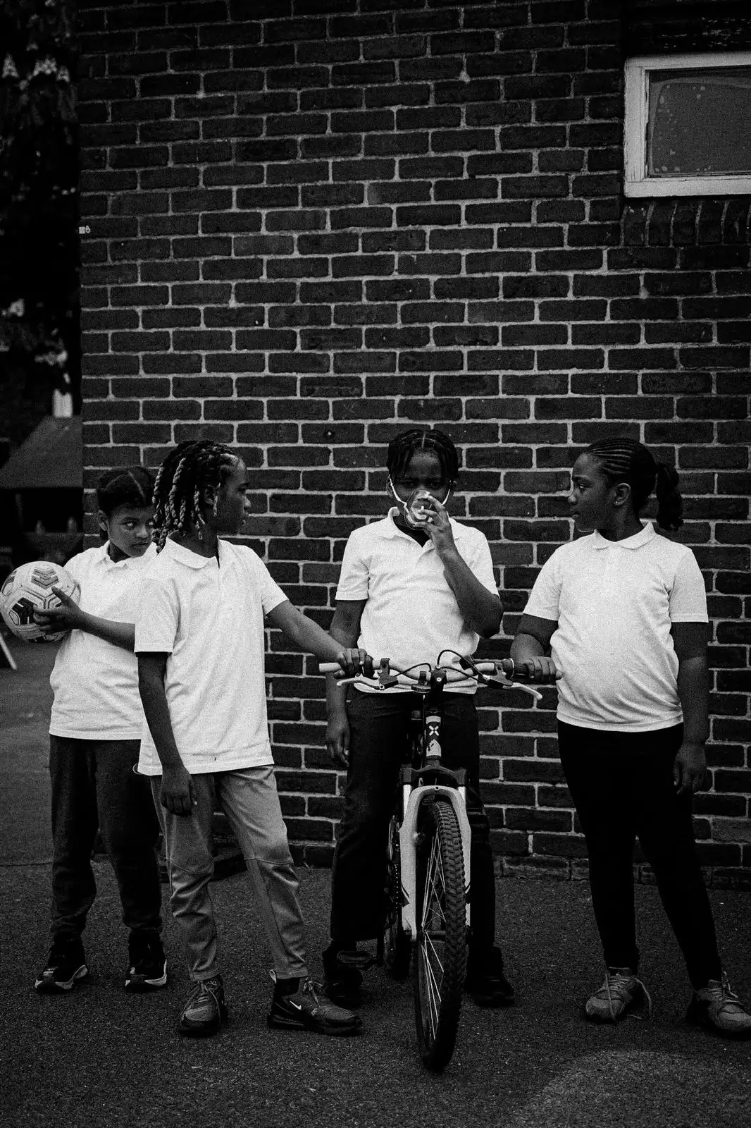 Black-and-white photograph of four children outside in front of a brick wall. One child is on a bike wearing a clear face mask – the other children are looking at them.