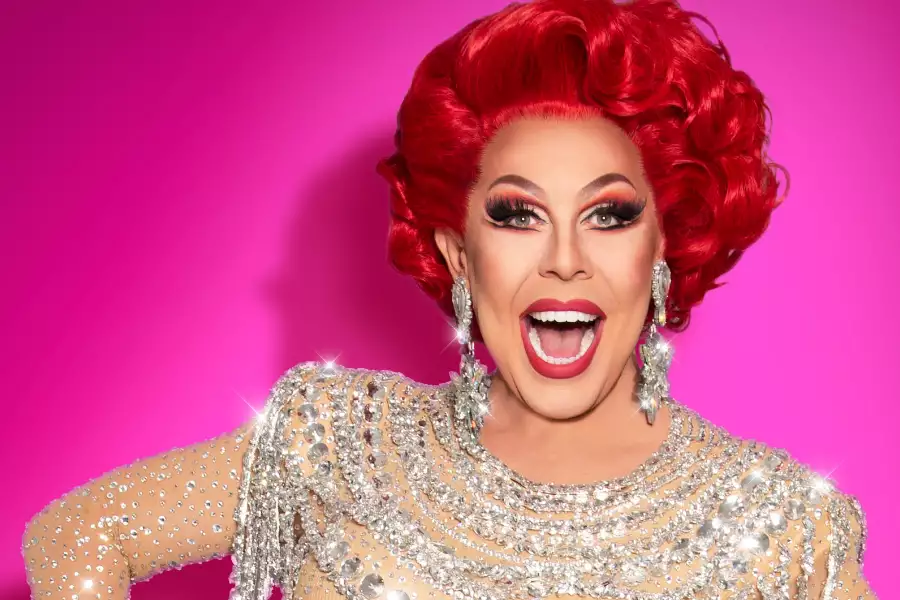 Drag queen La Voix poses with vibrant red hair, dramatic makeup, and a sparkling rhinestone against a bright pink background