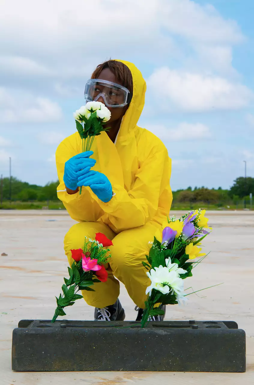 A person wearing a yellow hazmat suit, blue latex gloves and protective glasses. They are crouched on the floor outside with bunches of colourful flowers.