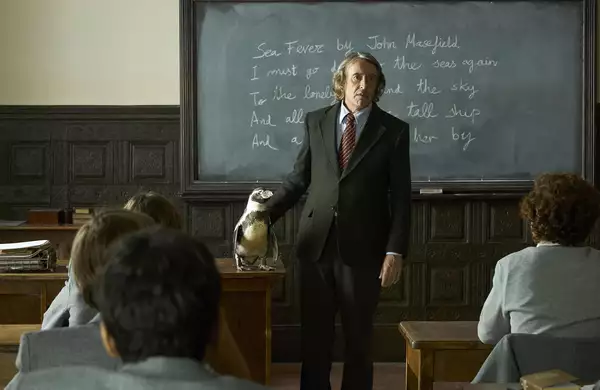A still from The Penguin Lessons showing a classroom scene with Steve Coogan. He is wearing a suit and standing in front of a blackboard. He rests one hand on a penguin standing on a wooden desk. Students in grey uniforms sit at wooden desks.