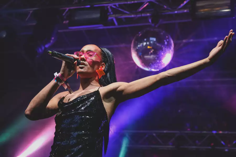A person singing on stage with a disco ball and purple lighting. They are wearing a black glittery dress and pink sunglasses.