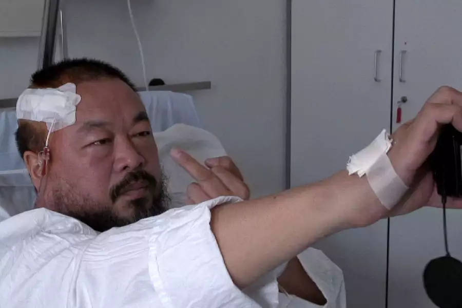 Ai Weiwei in a hospital bed with a bandage on his head, taking a photo of himself pulling a middle finger