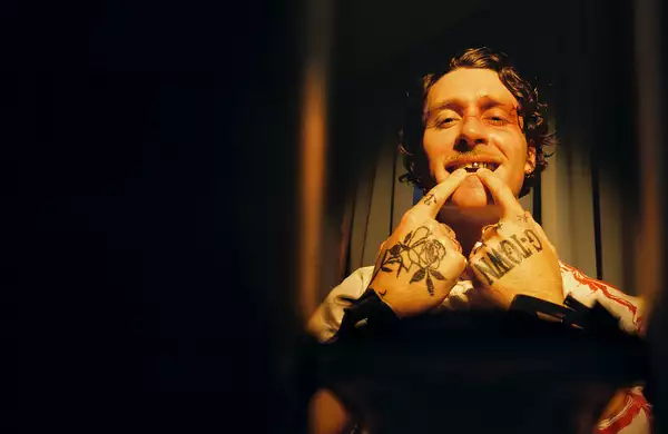 Photo of Kettama on a dark brown, shadowed background. His hands are tattooed and he is pointing at his metal teeth.