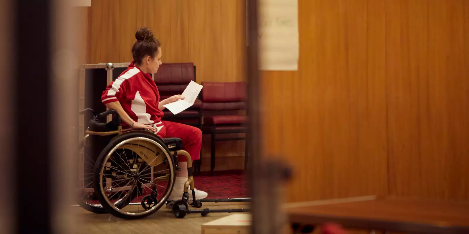 A person in a wheelchair reading from a sheet of paper in a wooden brown rehearsal room