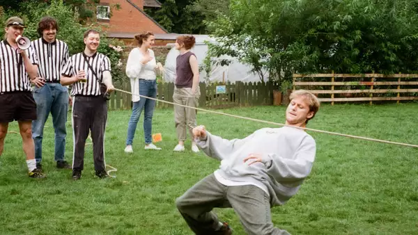 A person at Semi Peppered Sports Day in a grey sweatshirt plays limbo under a rope on a grassy lawn while others, including referees, watch and stand nearby.