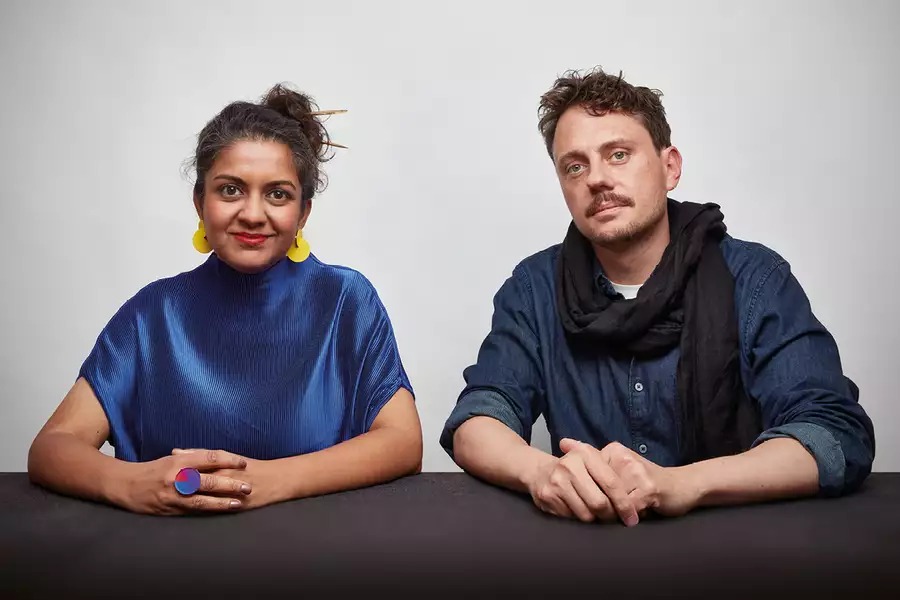 Two members of Superflux sat at a table, looking at the camera. The person on the left is wearing a shiny blue top and yellow earrings. The person on the right is wearing a blue shirt and black scarf.