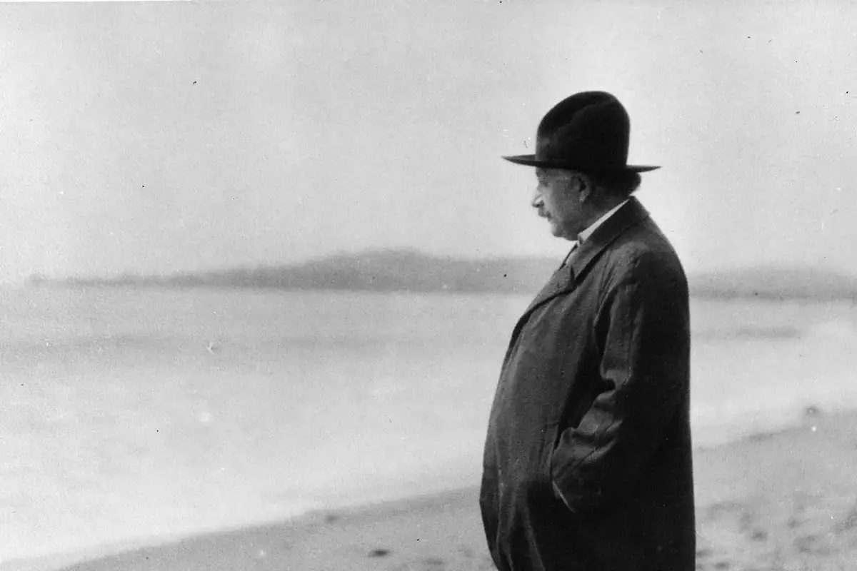 An archive image of Albert Einstein standing on a beach looking out towards the sea.
