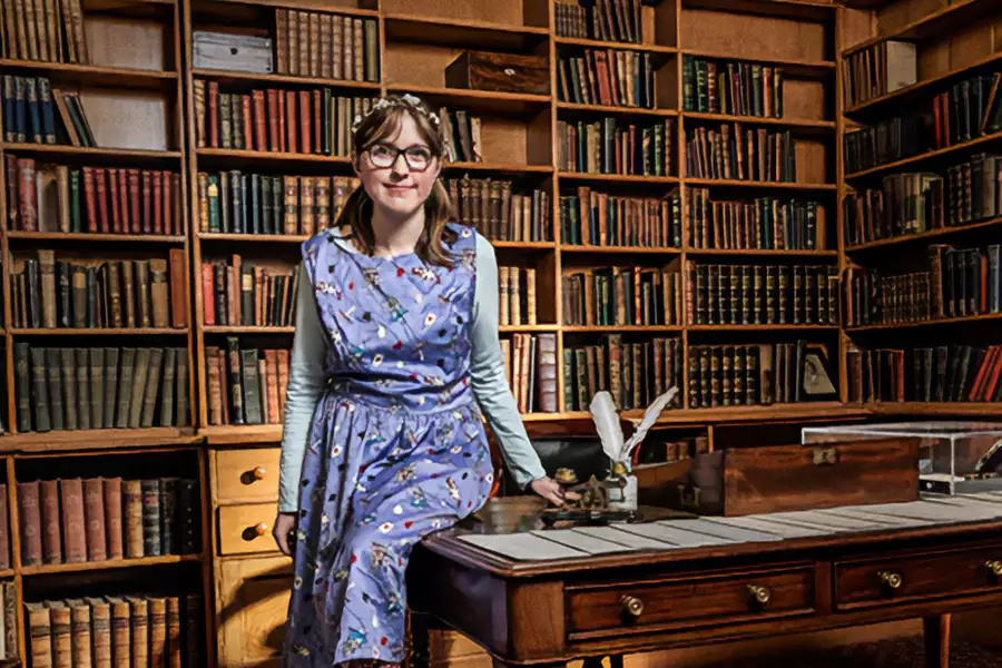 Sally Hirst wearing a blue patterned dress, smiling and perched on an old wood desk in a library.