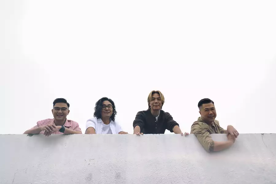 Four members of Rubberband leaning over a grey wall outside, smiling. The background is overcast.