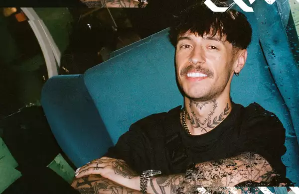 blk. smiling while sitting in a blue chair, wearing a black shirt and silver accessories.