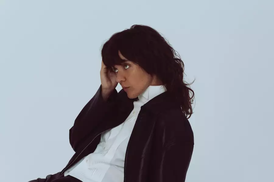 Courtney Barnett wearing a black jacket and a white shirt on a pale background