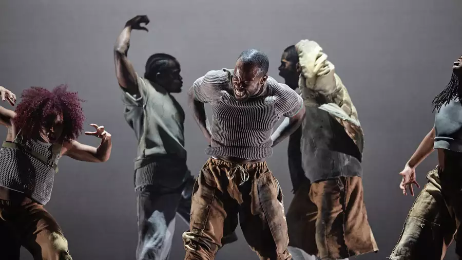 Five contemporary dancers perform with intense, expressive movements on stage, dressed in earthy-toned clothing. The central dancer leads the powerful scene, surrounded by others in dynamic, dramatic poses.