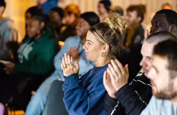 A room of people clapping. In the centre is a woman wearing a blue jumper clapping.