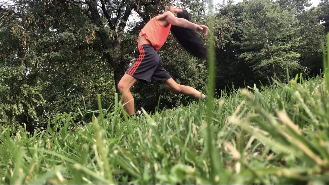 A diagonal photo of ​​Benedict Nguyễn, wearing black shorts, orange tank, and black and orange sneakers, lunging forward on left leg with back arched  left arm reaching behind her body, in a field with green grass and trees in background.
