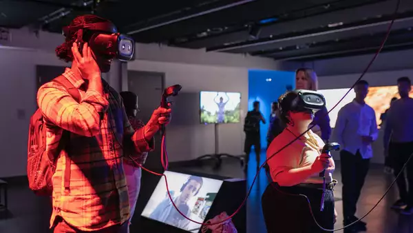 A man and a woman standing in a darkened art gallery, each wearing a Virtual Reality headset and holding a black controller. They are playing a game by the artist Rachel Maclean.