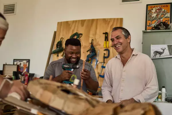 Two people smiling and laughing in a workshop setting for THE HERDS. One is holding a drill.