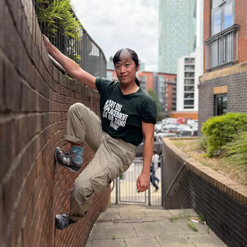 Louiseanne Wong hangs from a ledge with one arm and faces the camera. One foot is wedged more into the brick wall. There is a skyscraper behind them amongst other residential buildings.