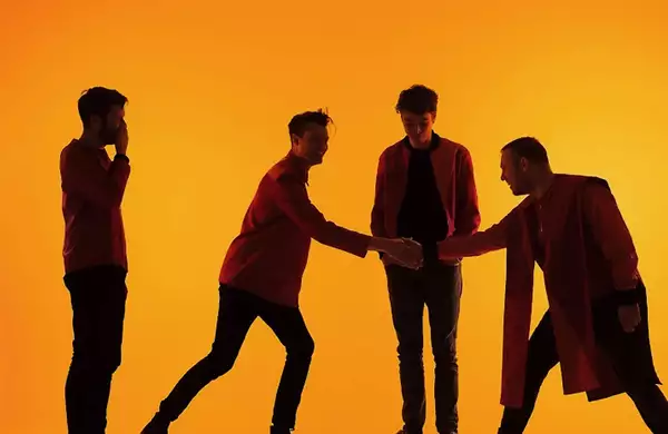 Four members of Everything Everything stand against a vibrant orange background, dressed in red and black, with two engaging in a handshake while the others observe. Their silhouettes and dynamic poses create a visually striking composition.