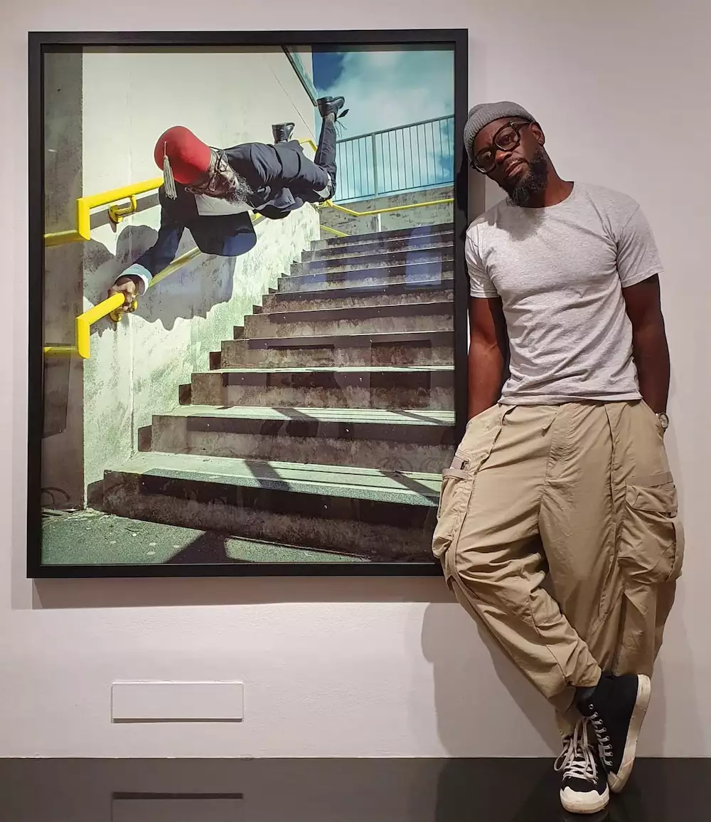 Benji Reid wearing beige trousers, a light grey top and a grey beanie. He is stood next to one of his photographs, which is framed on a white wall.