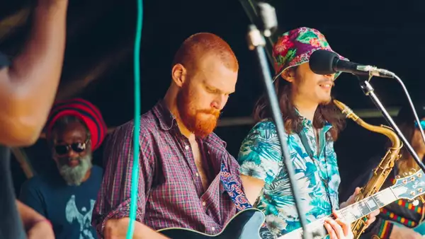 A group of people performing on stage. One holds a saxophone, another holds a guitar.