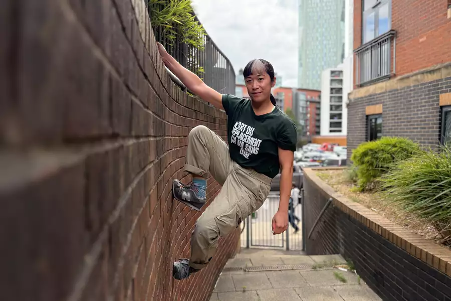 Louiseanne Wong hangs from a ledge with one arm and faces the camera. One foot is wedged more into the brick wall. There is a skyscraper behind them amongst other residential buildings.