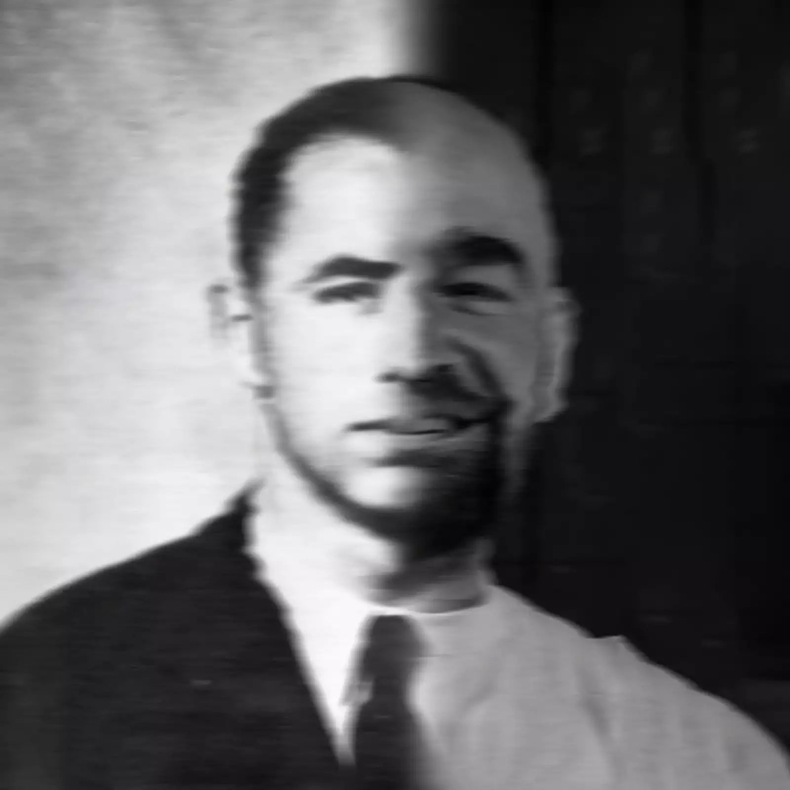 Black and white photo headshot of a person. One half of their body is clean-shaven and wearing a suit and tie; the other half is not.