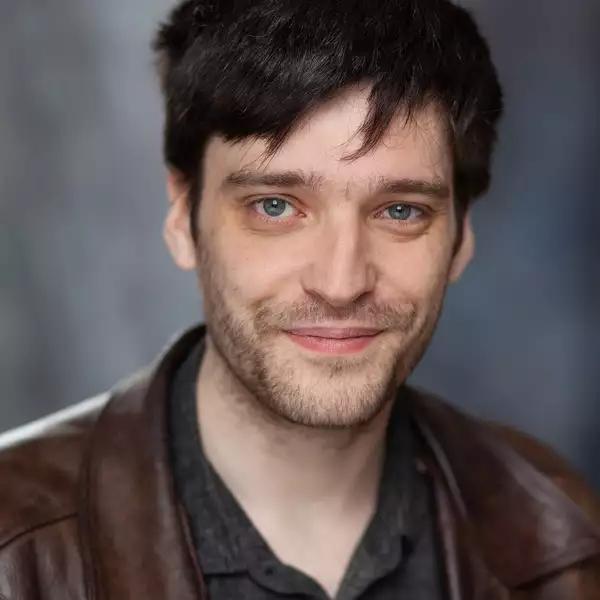Headshot of Elliot Hughes smiling and wearing a brown jacket