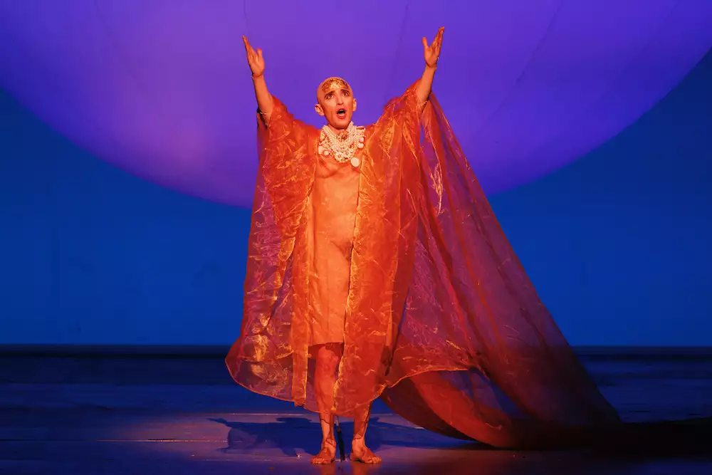 A person on stage singing with their arms in the air. They are wearing an orange, translucent cape. The background is blue.