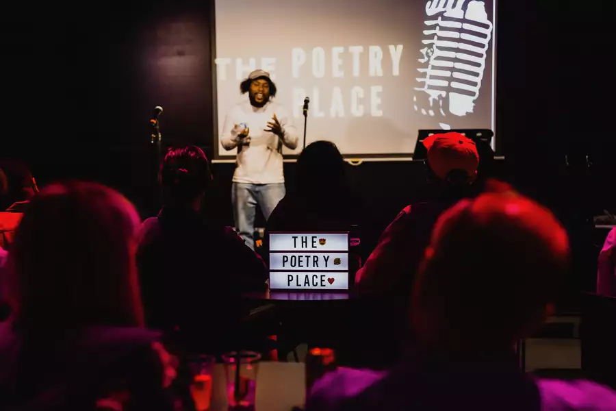 A performer is on stage in the Social with the words 'The Poetry Place' projected on the screen behind them. There is a lit-up sign that says 'The Poetry Place' in front of the stage. The room is dimly lit.