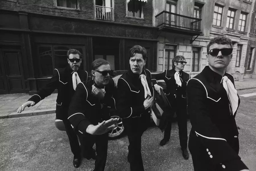 Black and white photograph of the five members of The Hives wearing matching suits and sunglasses striking dynamic poses on a city street. There is a car in the background.