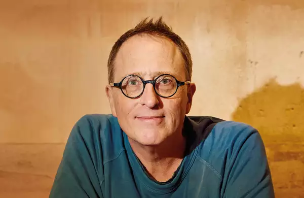 Jon Ronson smiling in front of a wall, wearing a blue t-shirt and glasses
