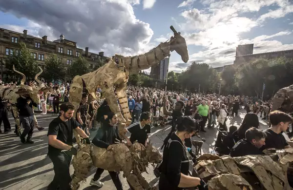 Performers parade life-sized cardboard animal puppets through a city square, watched by a large crowd on a sunny day.